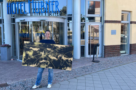 Künstlerin Jasmin Anderleit präsentiert mit einem Lächeln eines ihrer farbenfrohen Werke vor dem Strand-Hotel Hübner in Warnemünde.//Foto: Hotel Hübner