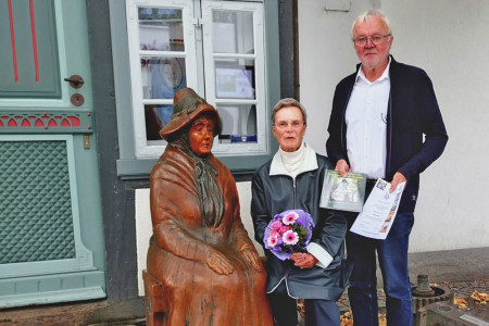 Die 10.000ste Museumsbesucherin 2025 Jutta Saedler und Werner Kreuscher vom Museumsverein posieren neben der von Harald Wroost geschnitzten Holzfigur „Die Warnemünderin“.//Foto: Hanne Hoppe