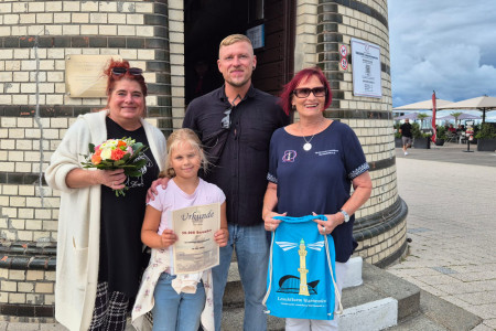 Die Rostocker Sch&uuml;lerin Ida Deuscher (2. v. l.) nutzt gemeinsam mit Oma Anna Spiller und Papa Paul Spiller die letzte Ferienwoche f&uuml;r einen Besuch des Warnem&uuml;nder Leuchtturms. Dort wurde sie von Inge Morenz vom Leuchtturmverein (r.) prompt als 50.000. Besucherin der Saison 2026 begr&uuml;&szlig;t.
