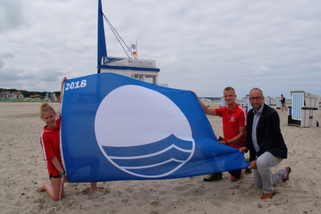 Sie hissen die &bdquo;Blaue Flagge&ldquo; am Strand von Warnem&uuml;nde: die Rettungsschwimmer der DRK-Wasserwacht Alexandra Sievert und Arian Baume und Tourismusdirektor Matthias Fromm (v.l.).