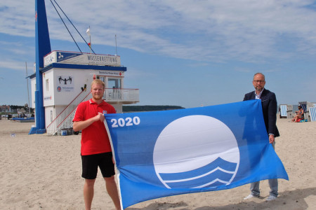Seit heute weht wieder die „Blaue Flagge“ an den Stränden von Warnemünde und Markgrafenheide. Eric Reck von der DRK Wasserwacht (l.) und Matthias Fromm hissten das international gültige Umweltsymbol am Strandaufgang 10.