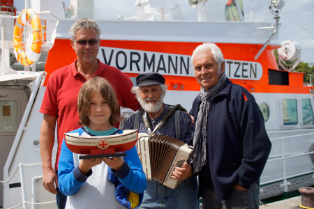 Jörg Westphal (l.) freut sich, die beiden Musiker Columbo (mitte) und Hein Schöler an Bord der "Vormann Jantzen" begrüßen zu dürfen. Mit dabei auch Schölers Enkel Hardy, der ein mit 90 Euro gefülltes Rettungsschiffchen an die Seenotretter übergibt.